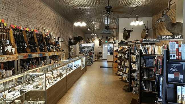 Inside Guns and Glory — firearms displayed along the left wall under taxidermy mounts, Christian Living book section labeled on the right wall. The two-aisle layout shows the shop's unique combination of gun shop and Christian bookstore.